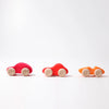 Three small Grimm's Colored Wooden Cars—pink, red, and orange—each with four light wheels, are lined up on a white surface. These non-toxic toys offer safe, colorful fun for little ones.