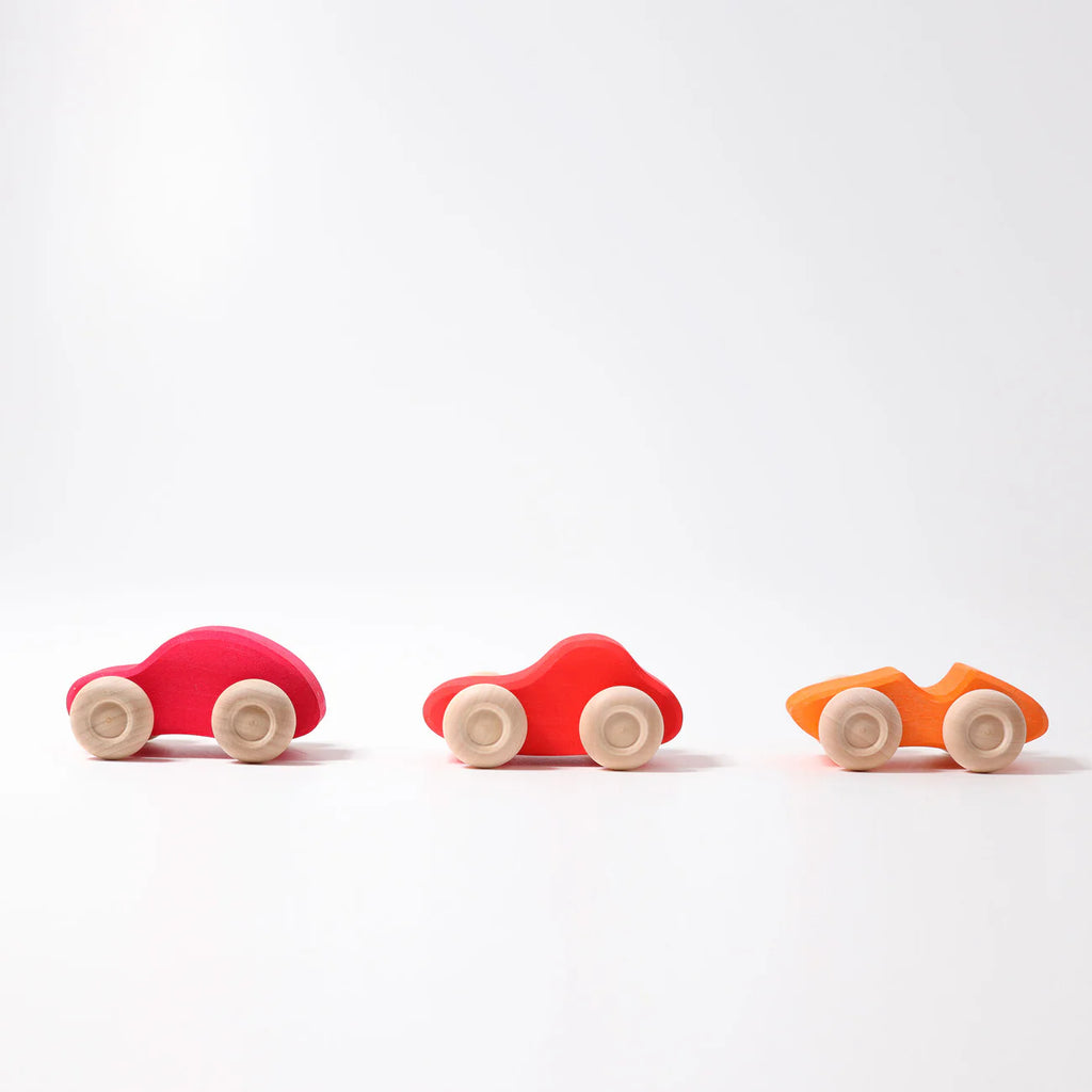 Three small Grimm's Colored Wooden Cars—pink, red, and orange—each with four light wheels, are lined up on a white surface. These non-toxic toys offer safe, colorful fun for little ones.