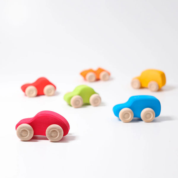Six Grimm's Colored Wooden Cars in red, blue, green, yellow, and orange sit on a white surface. These non-toxic wooden toys feature simple designs and natural wooden wheels for safe play.