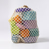 A mesh bag filled with colorful Grimm's Tree Slices Building Blocks against a plain white background. The non-toxic blocks come in various shades of purple, green, blue, and orange, and the netting allows for the vibrant colors to be visible through it. Perfectly safe for young children to explore and enjoy.