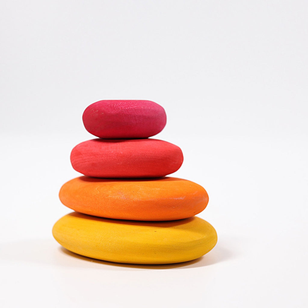 Four smooth, oval-shaped, organic stone shapes are stacked on top of each other on a white background. The stones, from bottom to top, are yellow, orange, red, and pink. Made from lime wood with a non-toxic color stain, each stone slightly varies in size, gradually getting smaller toward the top. These are the Grimm's Fire Pebbles Stacker.