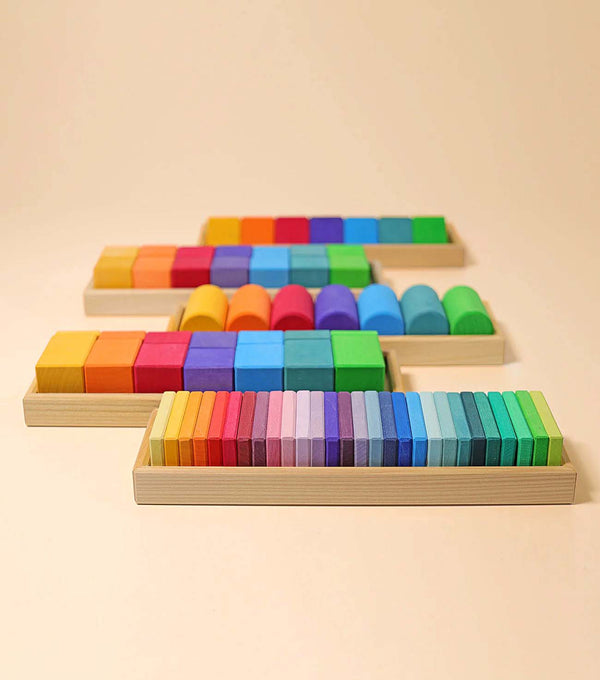 Grimm's Shapes and Colors Set features four wooden trays neatly displaying vibrant geometric building blocks in various shapes and sizes, arranged in color gradients, ideal for fine motor skill development against a light background.