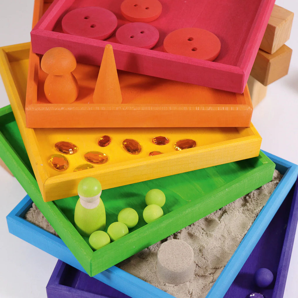 Grimm's Rainbow Frames (coming soon) create a playful kids workspace with colorful wooden trays, each filled with items like buttons, gems, and sand, arranged in rainbow order from red to purple on a white background.