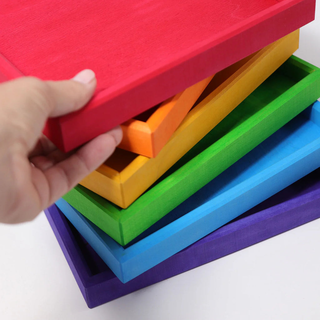 A hand pulls out Grimm's Rainbow Frames (coming soon): a stack of colorful wooden trays in red, orange, yellow, green, blue, and purple—perfect for brightening any kids workspace.
