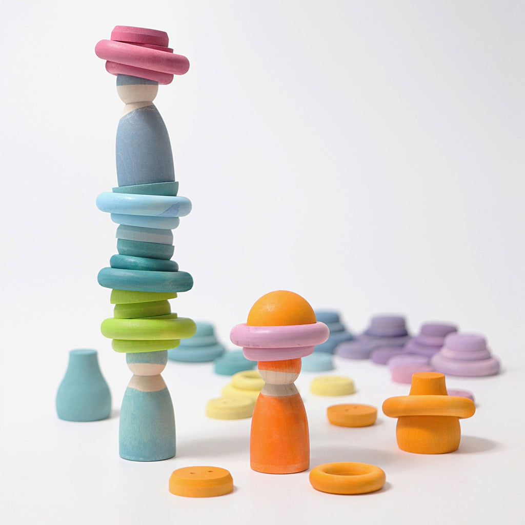 Grimm's Pastel Building Rings sit on a white surface, with wooden figurines adorned in colorful, stackable pieces of various shapes and pastel hues. In the background, additional stacking and sorting toys in shades of pink, purple, blue, and yellow are scattered; ideal for enhancing fine motor skills.