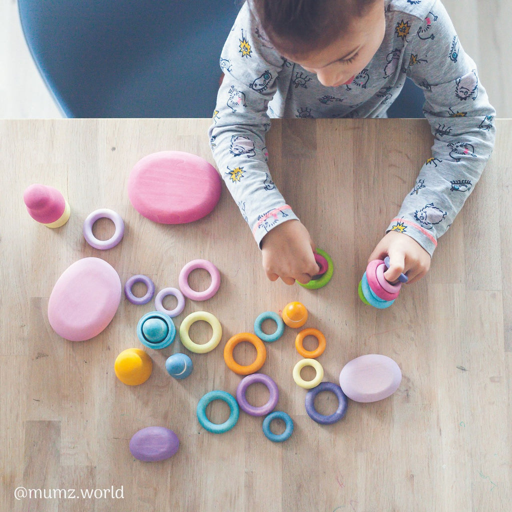 A child wearing a long-sleeve patterned shirt is sitting at a wooden table, arranging colorful Grimm's Pastel Building Rings along with other geometric shapes. The table is filled with stacking and sorting toys of various sizes and colors, such as pink, purple, blue, yellow, and orange, enhancing their fine motor skills.