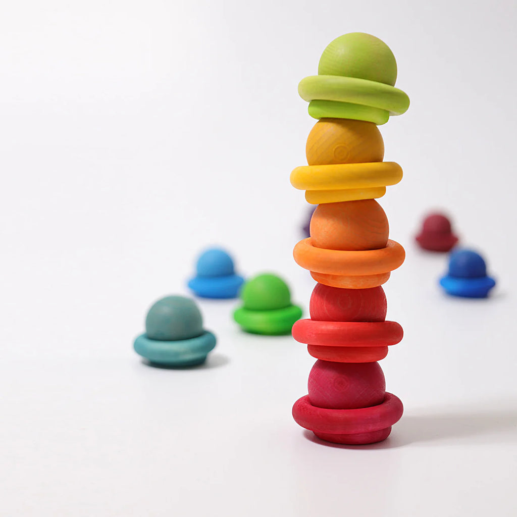 Grimm's Building Rings - Rainbow are stacked upright on a white surface, with vibrant rings and balls scattered in the background—perfect for developing children's fine motor skills.