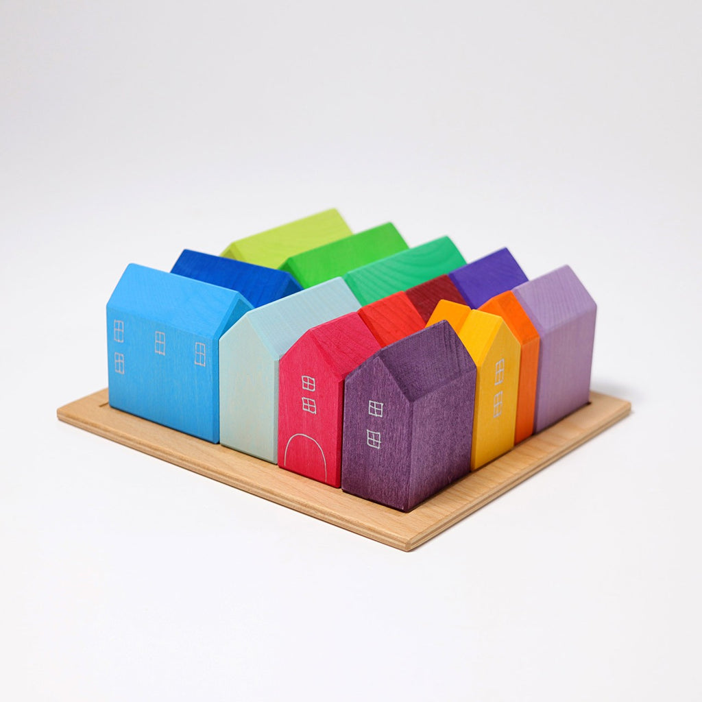 A set of vibrant, wooden, house-shaped blocks neatly positioned on a wooden base inspires the creativity of a city planner. Each block in Grimm's Houses is painted in distinct hues—blue, red, green, and purple—and features simple window and door details reminiscent of modern urban landscapes.