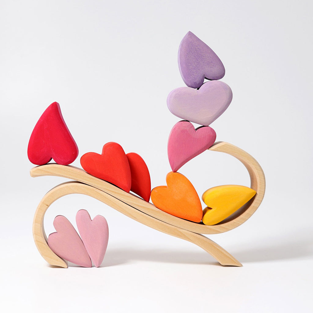 A whimsical wooden sculpture featuring Grimm's Red Hearts Building Blocks in shades of red, orange, yellow, pink, and purple, arranged in a curved, winding pattern on a light wood base. As a charming desk-toy enhancing fine motor skills, the hearts are stacked and balanced creatively, resembling an abstract tree.