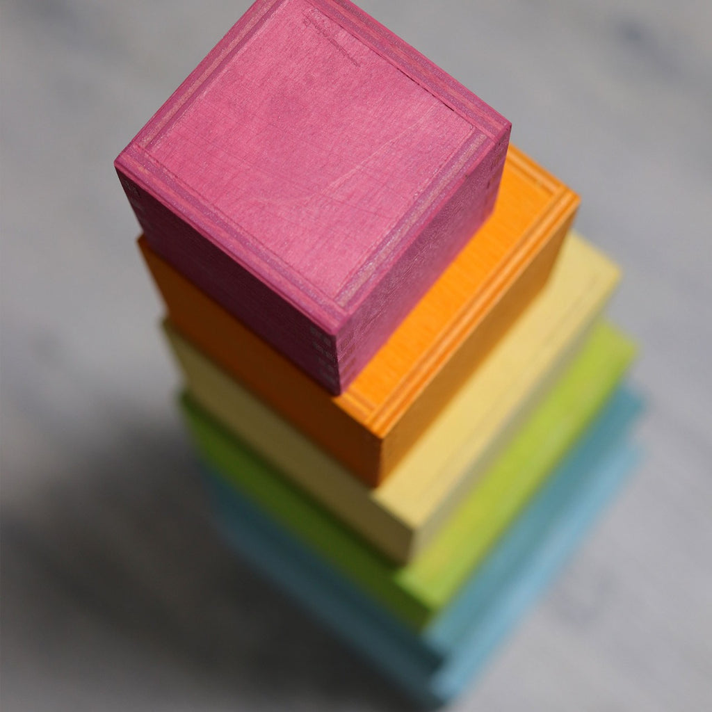 A vertical stack of non-toxic wooden blocks, each painted a different color: pink on top, followed by orange, yellow, green, and blue. The Grimm's Large Pastel Set of Boxes is arranged from largest at the bottom to smallest at the top, set against a neutral gray background.