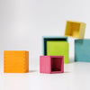 A versatile toy, the Grimm's Large Pastel Set of Boxes are scattered on a white surface in orange, pink, green, blue, and yellow. Some boxes are stacked while others are standing alone or tilted. Made from non-toxic materials, they're perfect for kids to stack and sort safely against a plain white background.