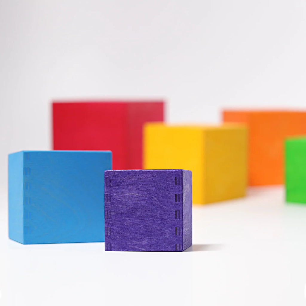 Grimm's Large Set of Boxes - Rainbow features six vibrant, non-toxic wooden boxes—blue, purple, red, yellow, orange, and green—stacked on a white surface. The purple box stands out in sharp focus at the center front.