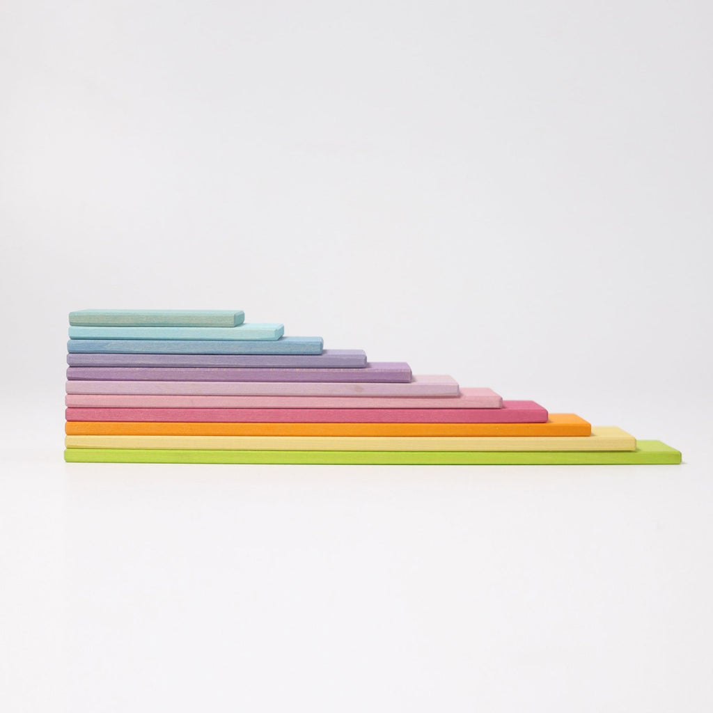 A tiered stack of colorful rectangular foam blocks arranged in ascending order from left to right. The smallest block is light blue, followed by purple, pink, orange, yellow, green, and a larger light green block at the top. This setup encourages open-ended play on a white background with the Grimm's Pastel Building Boards Set.