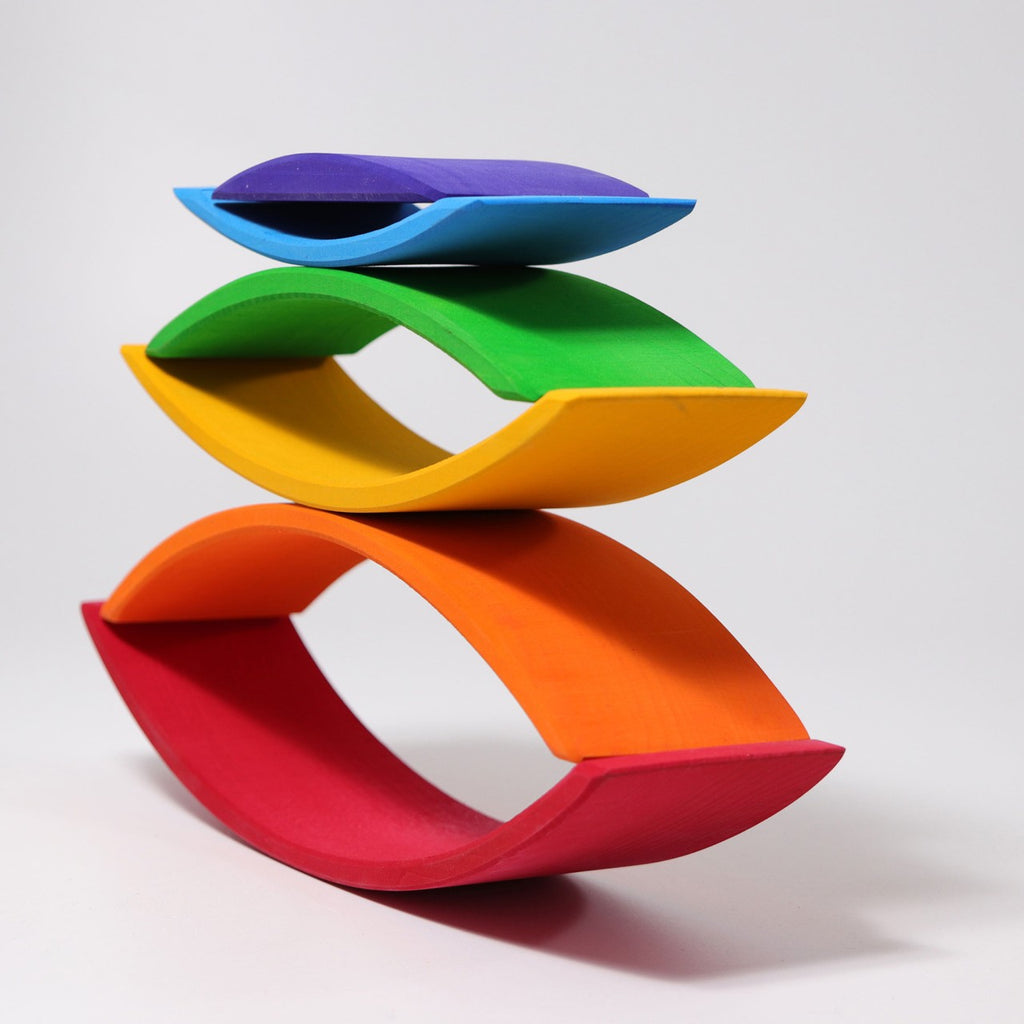 A stack of colorful, curved pieces of non-toxic wood, arranged in free-form balance. From bottom to top, the pieces are red, orange, yellow, green, and purple. The background is plain white, highlighting the colors and unique shapes reminiscent of Grimm's Rainbow Bridge.