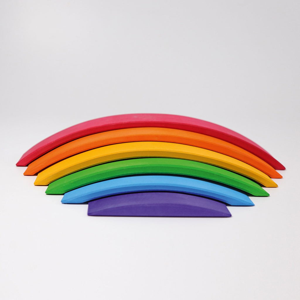 A set of colorful, curved wooden pieces are stacked in the shape of a Grimm's Rainbow Bridge on a light gray background. The non-toxic pieces are ordered from top to bottom in red, orange, yellow, green, blue, and purple.