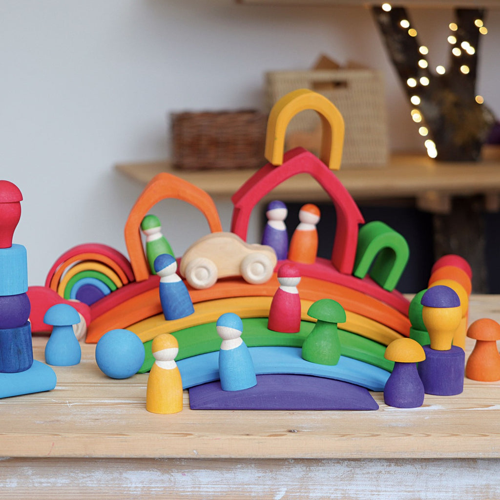 A playful wooden toy set featuring colorful arches, peg people, trees, and mushrooms arranged on a table. A small wooden car and Grimm's Rainbow Bridge in the background add charm. The non-toxic, vibrant pieces create a whimsical and inviting scene perfect for imaginative play.