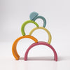 A minimalist arrangement of Grimm's Rainbow Pastel Stacker, stacked in a balanced structure, reminiscent of a rainbow tunnel. The non-toxic arches are colored in shades of blue, green, yellow, orange, and pink, set against a plain white background—perfect for toddlers to stack safely.