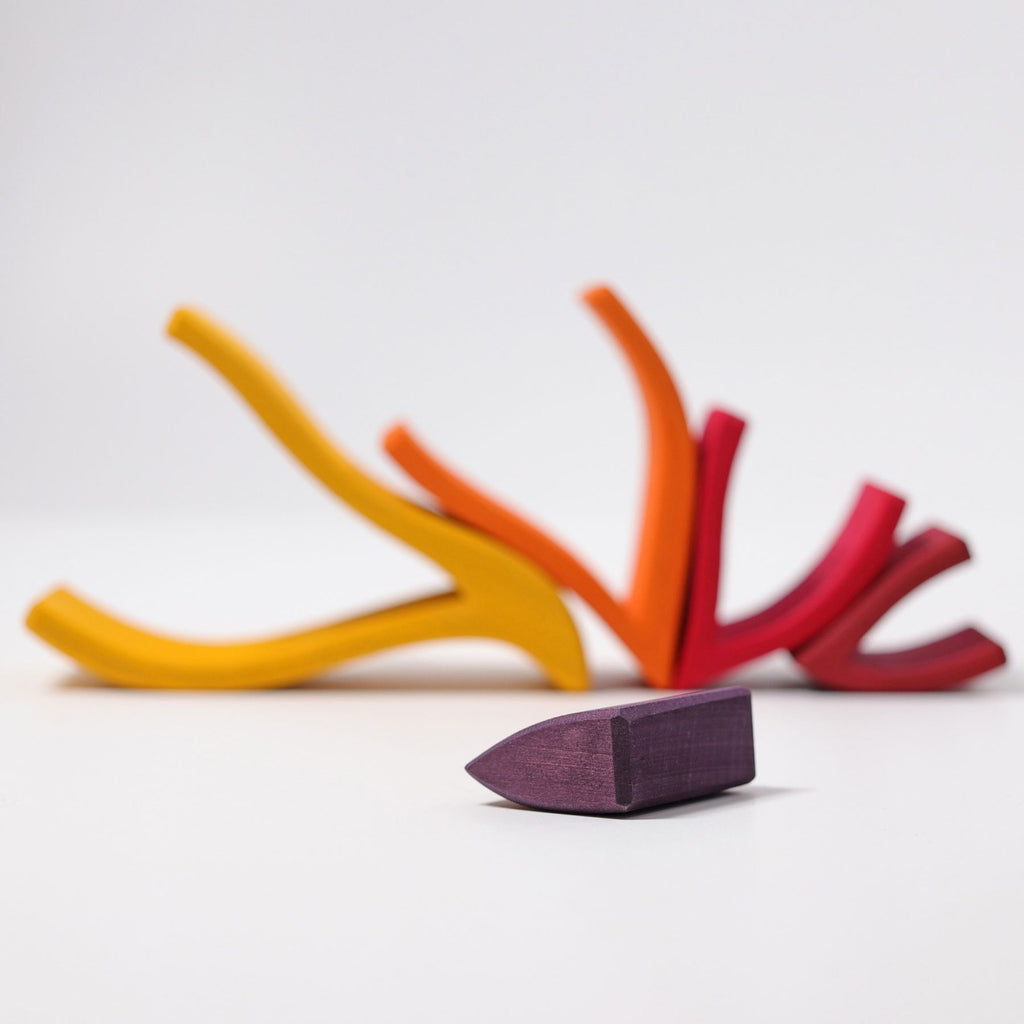 The Grimm's Fire Stacker consists of colorful, curved wooden pieces primarily in shades of yellow, orange, and red, arranged in an abstract, branching pattern against a white background. This charming wooden stacking toy also features a small purple piece shaped like a truncated cone placed in the foreground.