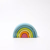 A Grimm's Small Wooden Rainbow, Pastel shaped like a rainbow tunnel, with five nested arches in descending size. From top to bottom, the versatile toy's arches are blue, light blue, green, yellow, and red. Made from non-toxic materials, the background is white and uncluttered.