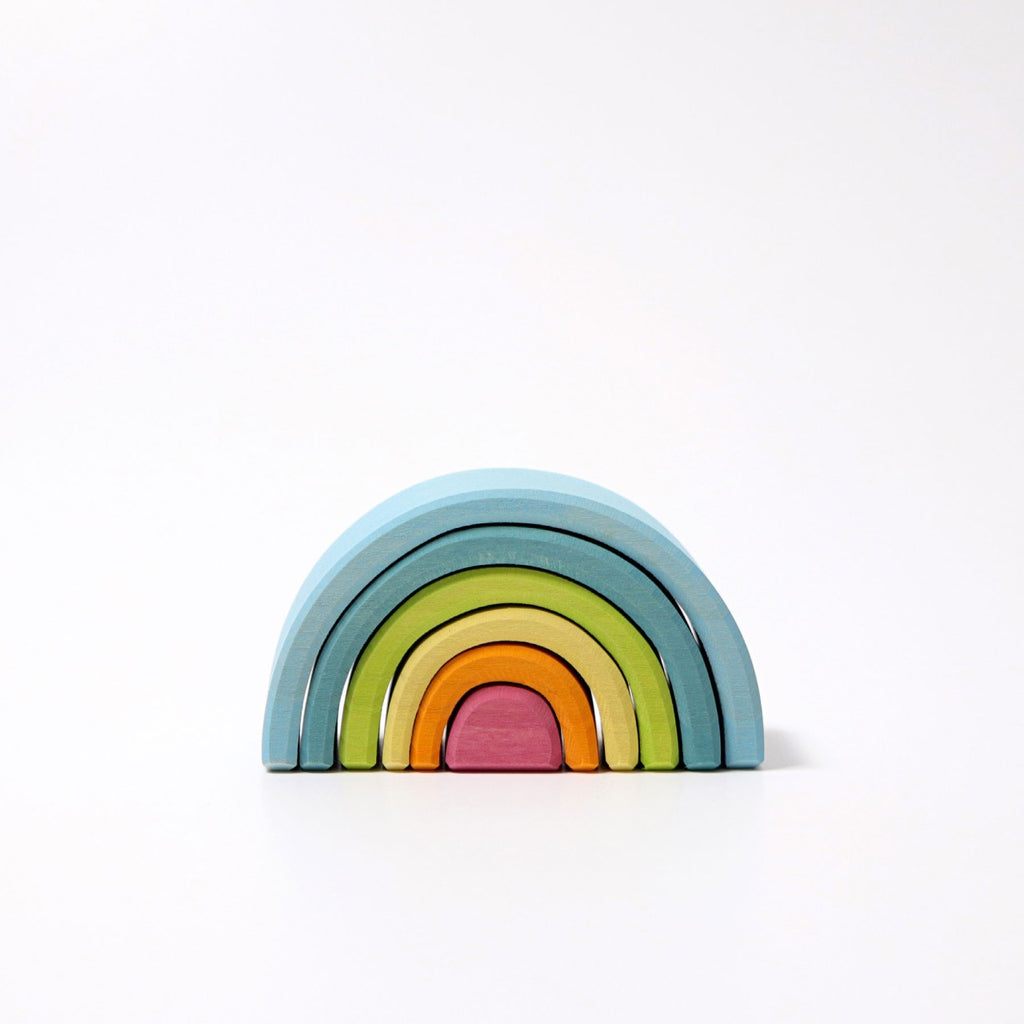 A Grimm's Small Wooden Rainbow, Pastel shaped like a rainbow tunnel, with five nested arches in descending size. From top to bottom, the versatile toy's arches are blue, light blue, green, yellow, and red. Made from non-toxic materials, the background is white and uncluttered.