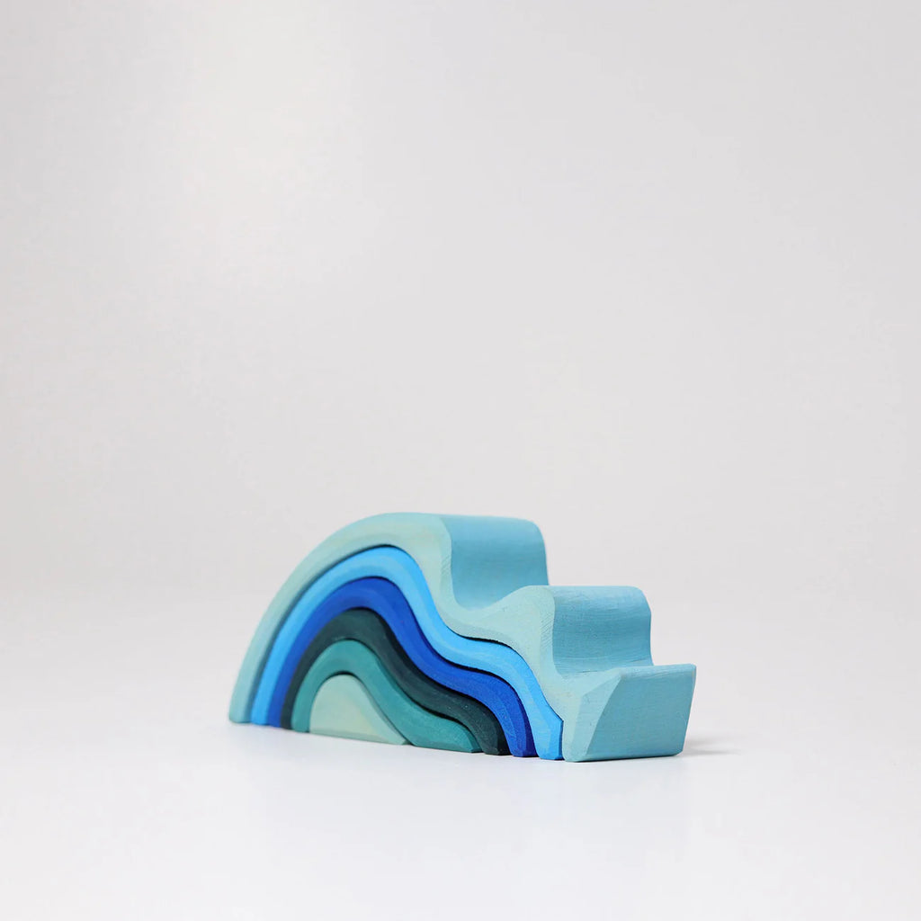 The Grimm's Ocean Waves, Small is a natural wood toddler stacking toy shaped like an abstract wave and painted in soothing blue shades, shown arranged on a white surface against a plain light background.