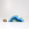 A seashell sits beside Grimm's Ocean Waves, Small—a natural wood toddler stacking toy featuring ocean wave shapes in blue and teal—set against a plain white background.