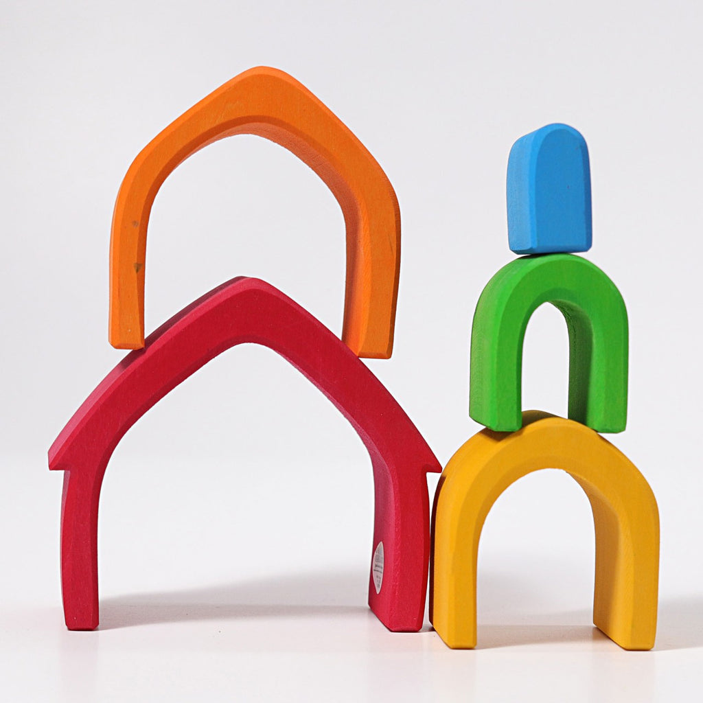 A colorful arrangement of Grimm's Colorful House Stacker stacked in a playful structure. The largest arch is red at the bottom left, topped by an orange arch, with green and yellow arches and a blue block forming a separate stack on the right against a white background—perfect for toddlers.