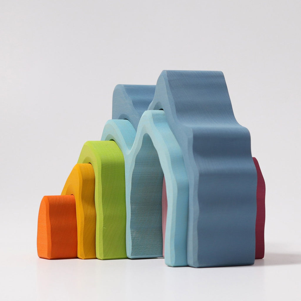 Grimm's Blue Cave Arch Stacking Toy features colorful, wavy wooden arches—orange, yellow, green, light blue, blue, and red—nested in rainbow order. Each piece is finished with natural oil and displayed on a plain white background.