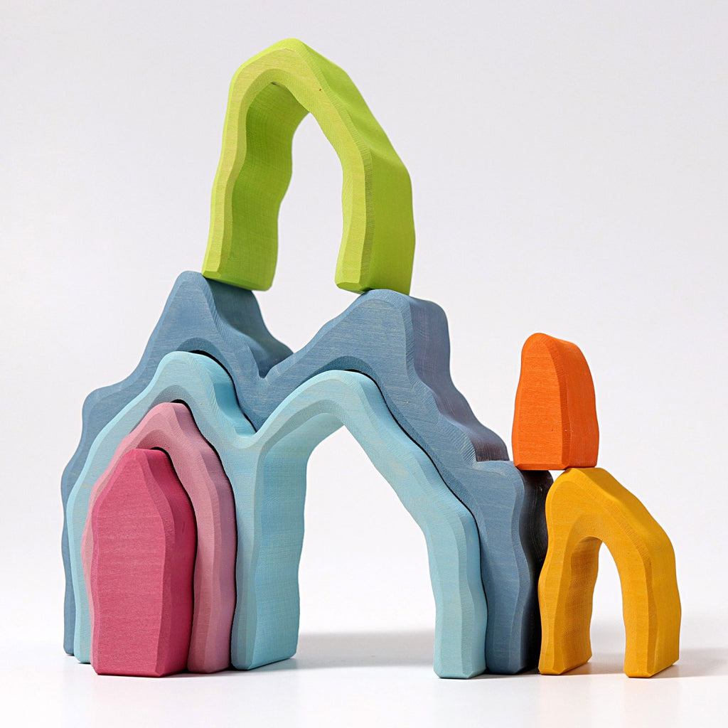 The Grimm's Blue Cave Arch Stacking Toy features colorful wooden blocks in cave arch shapes, arranged as abstract mountains and arches in blue, green, pink, orange, yellow, and red on a plain white background.