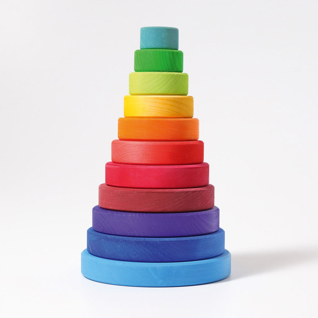 A variety of colorful wooden discs, available in blue, green, yellow, orange, and purple and of different sizes, are neatly arranged on a flat surface with a vertical rod at the center. The arrangement forms a Grimm's Stacking Tower in progress. This engaging toy enhances eye-hand coordination and fine motor skills. The background is white.