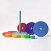 A variety of colorful wooden discs, available in blue, green, yellow, orange, and purple and of different sizes, are neatly arranged on a flat surface with a vertical rod at the center. The arrangement forms a Grimm's Stacking Tower in progress. This engaging toy enhances eye-hand coordination and fine motor skills. The background is white.