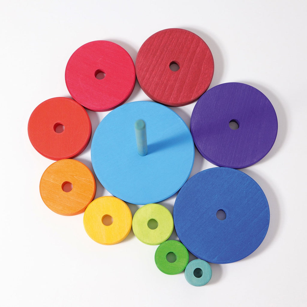 The Grimm's Stacking Tower features colorful, circular wooden discs varying in size, organized by color: red, orange, yellow, green, blue, purple, and natural wood. Placed next to a blue vertical rod, it’s designed to enhance eye-hand coordination and fine motor skills in children through playful learning.