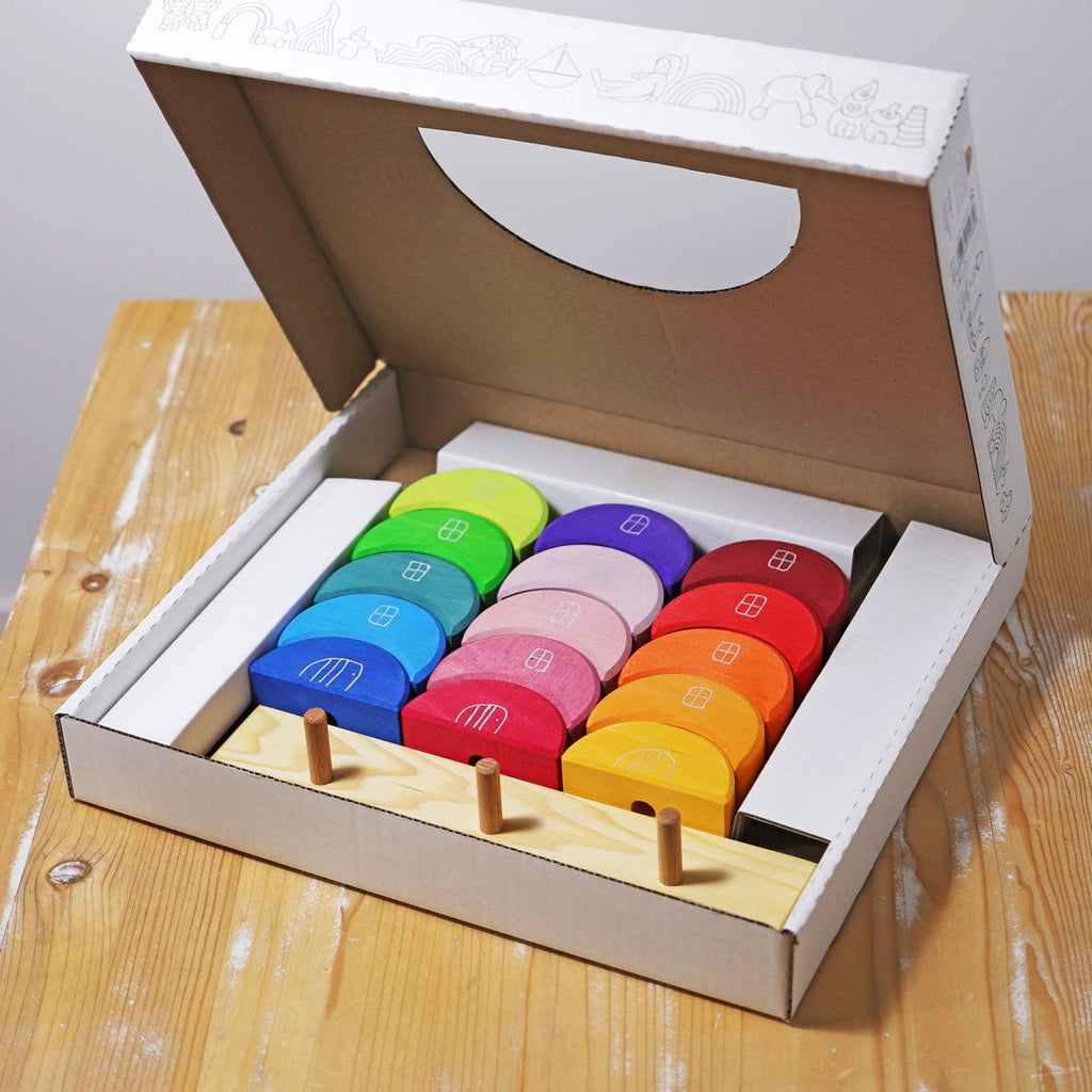 The cardboard box of Grimm's Moon Houses contains vibrant wooden blocks designed in house shapes, ideal for sorting and stacking activities. These blocks are neatly arranged on a wooden base with pegs, enhancing fine motor skills and eye-hand coordination. The inside lid of the box is adorned with decorative line drawings.