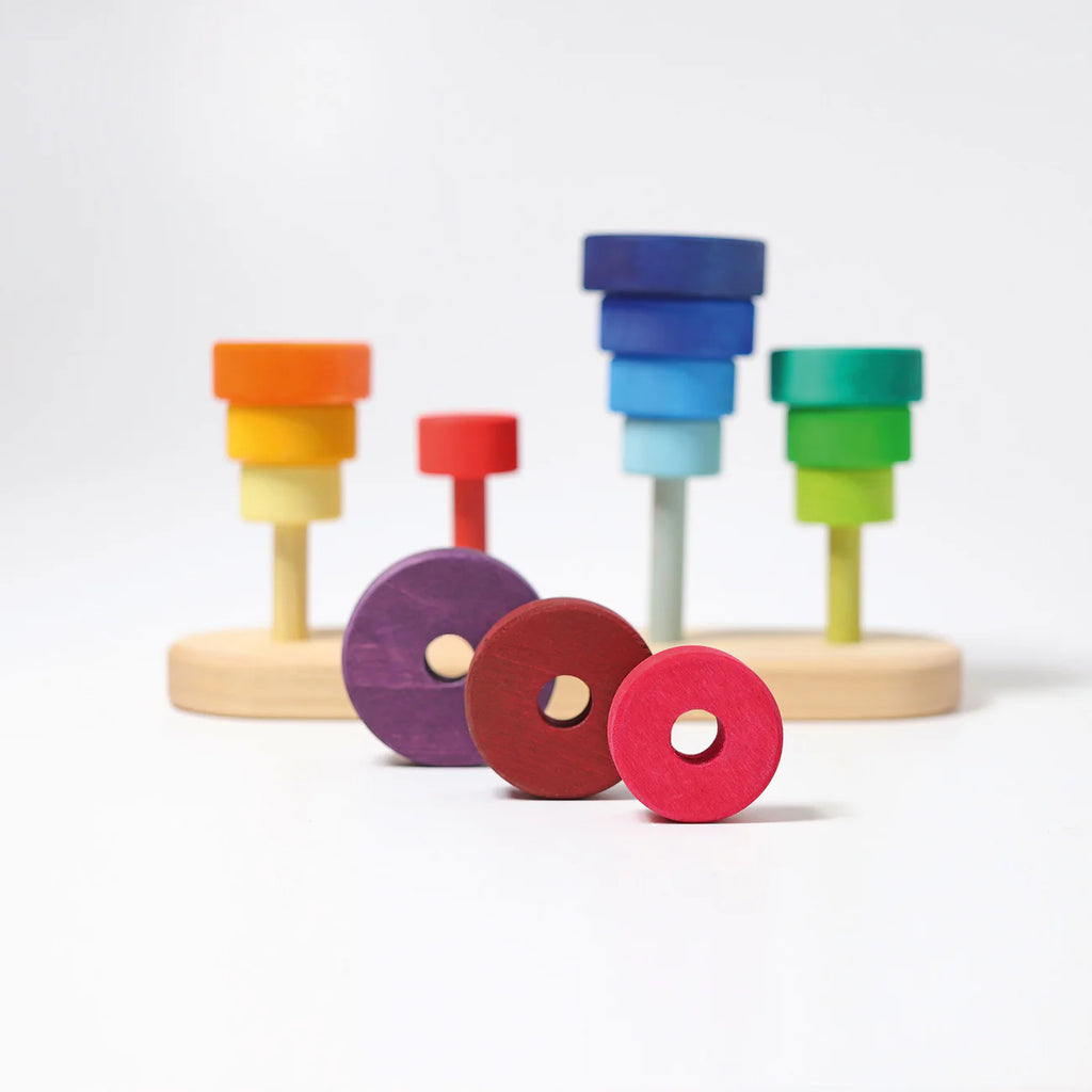 Grimm's Fabuto Stacker: A colorful wooden stacking toy shaped like trees, with rings of various sizes and colors on pegs, plus three loose discs (red, maroon, purple) in the foreground—all inspired by Grimms' signature color coordination.