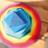 A top-down view of a stacked, multicolored Grimm's Giant Geometrical Stacking Tower. The pieces are triangular and arranged in a pyramid shape, creating a layered effect with vibrant colors including blue, green, yellow, and red. Perfect for developing fine motor skills, the non-toxic toy sits on blurred wooden planks.