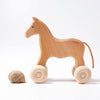 A Grimm's Wooden Horse Willy Push Toy, made in Germany, stands next to a seashell on a white background. The horse has a simple, minimalist design with a small tail made of string and is crafted from non-toxic materials.