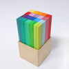 A wooden box holds a neat stack of colorful rectangular blocks, perfect for children's fine motor skills development. Arranged in a gradient, the blocks transition from shades of blue and green at the front to warmer hues of red, orange, and yellow towards the back. The background is plain white. The product is Grimm's Leonardo da Vinci Building Sticks.