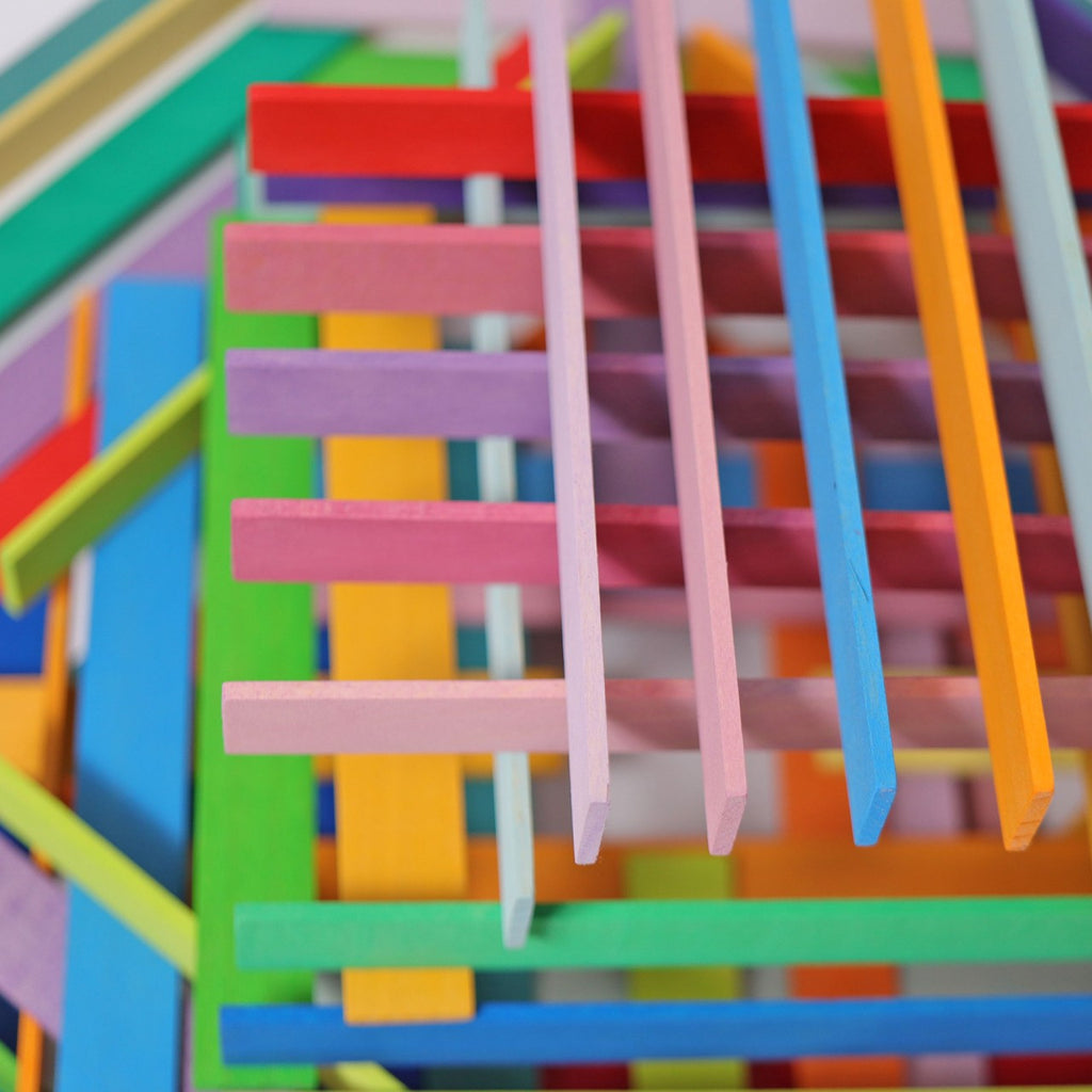 Colorful Grimm's Leonardo da Vinci Building Sticks resembling a children's building toy are arranged in a crisscross pattern, creating a visually intricate and playful composition. The arrangement, akin to Leonardo da Vinci constructions, features varied shades overlapping in a seemingly random yet cohesive manner.