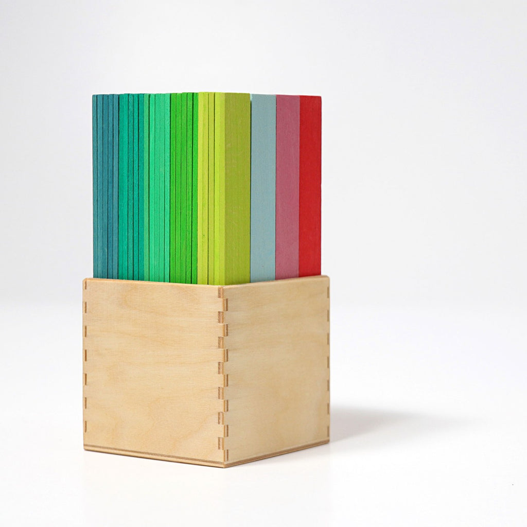 A wooden box with vertical multicolored wooden slats, perfect for Grimm's Leonardo da Vinci Building Sticks, arranged neatly inside. The slats are colored in various shades of blue, green, yellow, red, and gray. The box has dovetail joints and sits on a plain white surface against a white background.