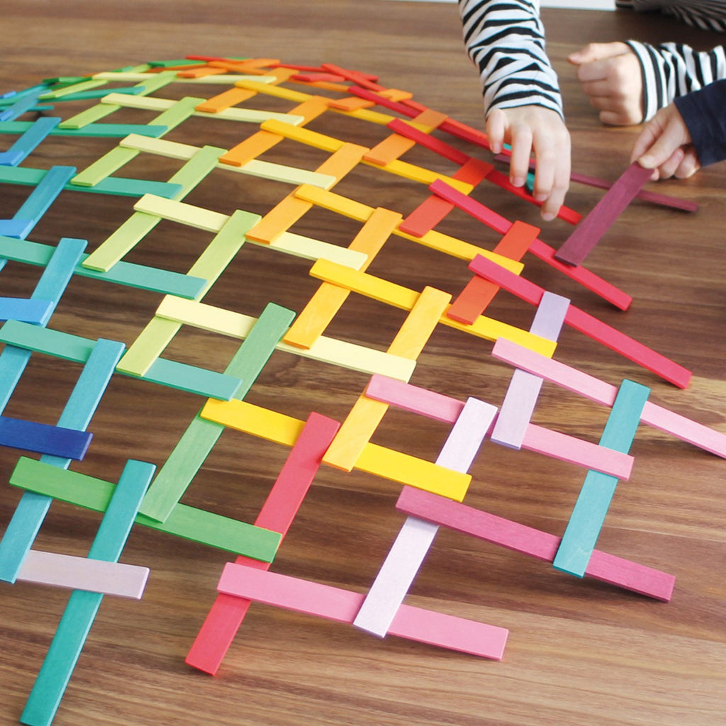 Two children are playing with a colorful, intricate pattern made of Grimm's Leonardo da Vinci Building Sticks on a wooden table. The sticks are arranged in an interlocking grid, creating a vibrant rainbow design. One child wears a striped shirt, and both have outstretched hands adjusting the pieces, honing their fine motor skills.