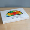 A white box of Grimm's Color Charts-Rally rests on a light wood surface, displaying vibrant stained wood tiles through its window—perfect for sparking creativity and building fine motor skills.