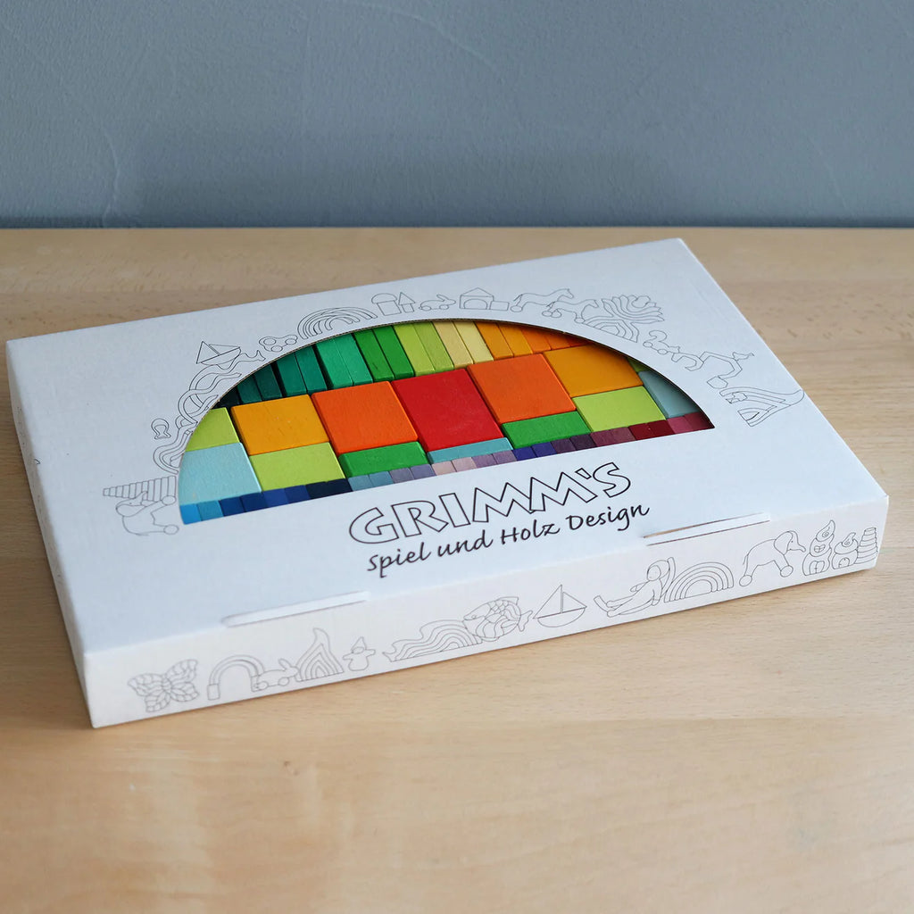 A white box of Grimm's Color Charts-Rally rests on a light wood surface, displaying vibrant stained wood tiles through its window—perfect for sparking creativity and building fine motor skills.