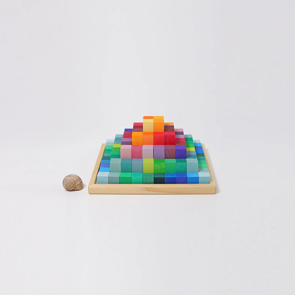 The Grimm's Stepped Pyramid Block Set - Small features colorful wooden blocks arranged in a gradient on a square tray, with a small brown snail shell to the left, all displayed on a white background.