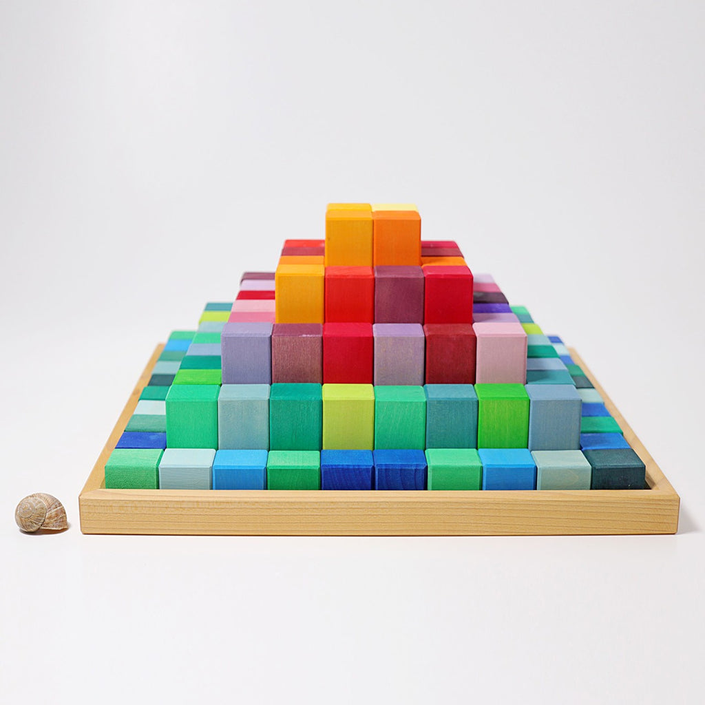 A Large Stepped Pyramid Block Set built from 100 colorful, non-toxic wood blocks arranged by size and color with a smaller, similarly patterned set beside it. The base blocks are shades of blue and green, transitioning to reds and oranges at the top. A small seashell sits on the left side, encouraging fine motor skills development.