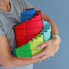 A person in a grey shirt holds several Grimm's Counterrotating Stepped Spiral blocks in vibrant red, green, and blue against a blue background. These colorful wooden toys help develop fine motor skills.