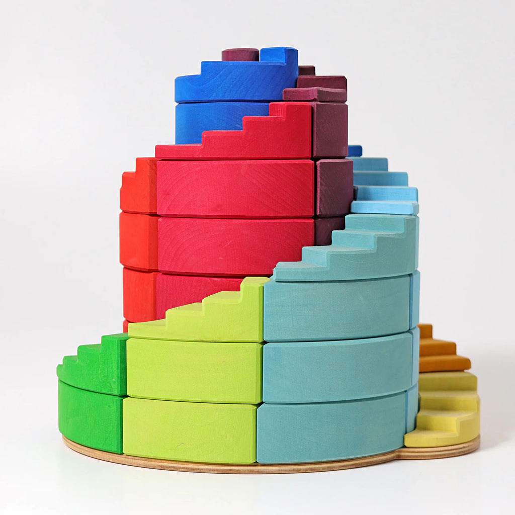 Grimm's Counterrotating Stepped Spiral features wooden blocks in a stepped spiral, arranged by color from green and yellow at the base to blue, red, and purple on top. Set against a white background, it’s ideal for building fine motor skills.