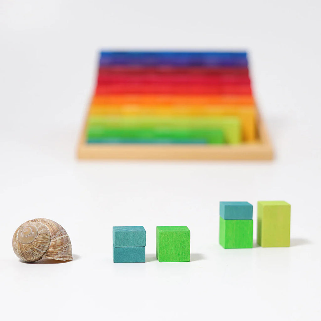 A brown snail shell rests next to pairs of blue and green Grimm's Stepped Counting Blocks - Small, with a blurred tray of rainbow blocks in the background on a white surface.