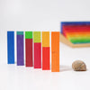 Six colorful rectangular Grimm's Stepped Counting Blocks - Small (coming soon) stand upright in a height- and color-graded row from blue to orange, with a snail shell on the right and more rainbow blocks blurred in the background.
