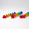 Small, colorful wooden shapes, including hearts, stars, and fish, are scattered on a white surface for a Grimm’s Sorting Game Rainbow Bowls with Tweezers game.