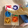 A toddler uses the wooden tweezers from a Grimm’s Sorting Game Rainbow Bowls with Tweezers set to sort wooden shapes into matching bowls, building fine motor skills.